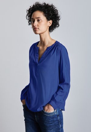 Blå langærmet bluse med V-hals og samlede skuldre, lavet af et glat stof. Kombineret med blå jeans, der viser et afslappet look.