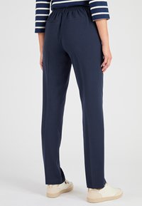 Donna che indossa pantaloni a gamba dritta blu navy, una maglietta a righe blu e bianca a maniche lunghe e sneakers bianche, in piedi di fronte a uno sfondo bianco.