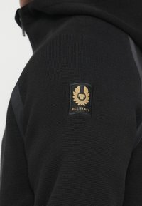 Μαύρο φούτερ με υφή και φερμουάρ, με χρυσή ετικέτα με το λογότυπο "Belstaff" στον ώμο. Ομαλή υφασμάτινη λεπτομέρεια στα πλαϊνά.