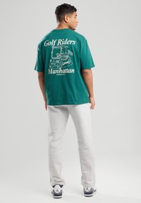 Vihreä puuvillapaita, jossa on golfkärrykuva ja teksti "Golf Riders Manhattan" painettuna selkäpuolelle, yhdistettynä harmaisiin collegehousuihin.