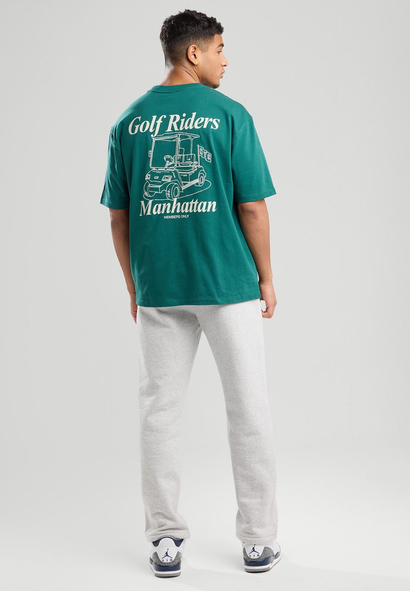Vihreä puuvillapaita, jossa on golfkärrykuva ja teksti "Golf Riders Manhattan" painettuna selkäpuolelle, yhdistettynä harmaisiin collegehousuihin.