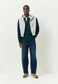 Zöld, hosszúujjú póló világosszürke kapucnis pulóver alatt, kék farmernadrággal és bézs sportcipővel, amelyeken türkiz kiegészítők találhatóak. Egyszerű, laza öltözet.