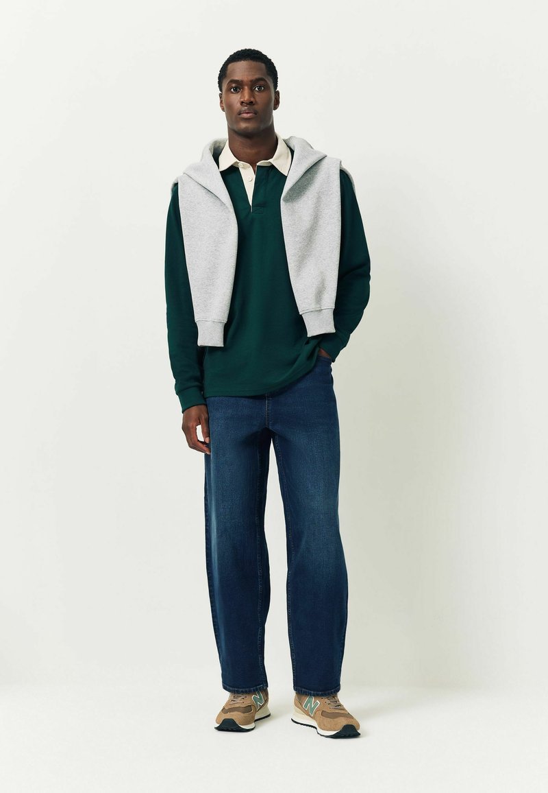 Zöld, hosszúujjú póló világosszürke kapucnis pulóver alatt, kék farmernadrággal és bézs sportcipővel, amelyeken türkiz kiegészítők találhatóak. Egyszerű, laza öltözet.