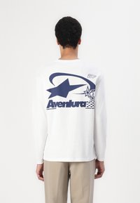 Hvit langermet t-skjorte med blå grafisk design med stjerner, med teksten "Aventura" på baksiden. Laget av bomull med avslappet passform.
