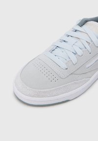 Ljusgrå mockasneaker med perforerad tå, vit sula, matchande ljusgrå skosnören och synlig Reebok-logotyp på sidan och tungan.