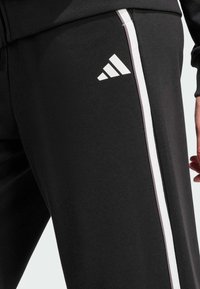 Musta urheiluhousut, jotka on valmistettu sileästä kankaasta. Niissä on valkoinen Adidas-logo ja kontrastiväriset sivuraitat vaaleammassa sävyssä.