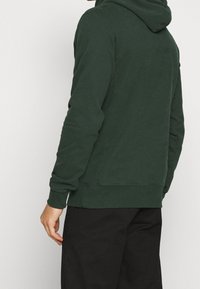 Persona con sudadera verde oscura y pantalones negros, vista desde atrás contra un fondo claro y liso.