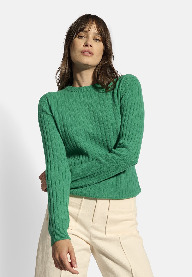 Jersey de punto verde acanalado con mangas largas y escote redondo, con un acabado texturizado y una cintura ajustada. Llevado con pantalones crema.