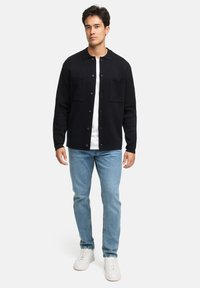 Sort, button-up jakke med krave og to frontlommer, båret over en hvid skjorte. Sammen med lyseblå jeans og hvide sneakers.