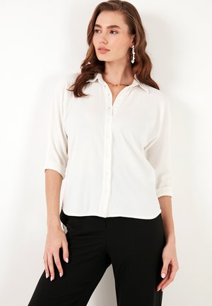 White button-up shirt with a textured finish, featuring a collar and rolled sleeves. Paired with black pants for a contrasting look.