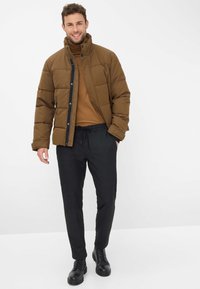 Bruine pufferjack met een hoge kraag, zwarte rits en een doorstikt ontwerp, gecombineerd met een bruine top en donkere broeken, afgewerkt met zwarte laarzen.