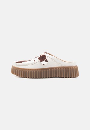 Scarpa bianca slip-on con stampa maculata marrone sulla parte superiore, suola spessa e scanalata marrone, e cuciture in stile mocassino, mostrata di lato su uno sfondo bianco.