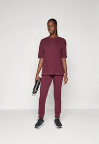 Bordeauxrotes Sportoutfit bestehend aus einem lockeren T-Shirt mit kurzen Ärmeln und enger zulaufender Hose. Das Modell hält eine schwarze Wasserflasche und trägt schwarze Turnschuhe.