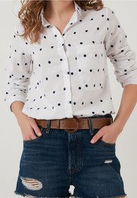Femme portant une chemise blanche à manches longues avec des pois noirs, rentrée dans un short en jean déchiré avec une ceinture marron, mains dans les poches.