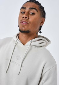 Sudadera gris claro con capucha con cordones, logotipo bordado en el pecho, textura suave y corte relajado. Accesorios plateados en los cordones.