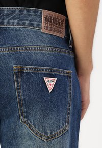 Denim jeans met een bruine leren patch op de taille met een ruitpatroon en een driehoekige rood-witte logo label op de achterzak.