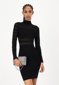 Svart bodycon-kjole med lange, gjennomsiktige stripete ermer og høy hals. Kombinert med en sølvfarget, pyntet clutch med kjedehank.