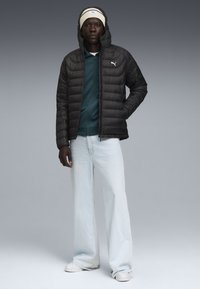 Zwarte pufferjack met ritssluiting en kap; gecombineerd met een groene ritssluiting top, lichtblauwe wijde jeans en witte sneakers.