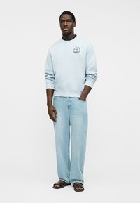 Hombre con sudadera azul claro, jeans azul claro holgados y sandalias oscuras, de pie con una mano en el bolsillo sobre fondo blanco.