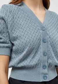 Světle modrý kardigan s texturovaným diamantovým vzorem, výstřihem do V, krátkými rukávy a pěti modrými knoflíky vpředu. Pletená tkanina.