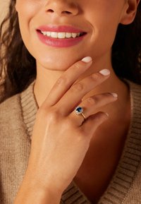 Gold ring featuring a central blue gemstone surrounded by smaller clear stones. Model's hand rests on her face, showcasing light nail polish.
