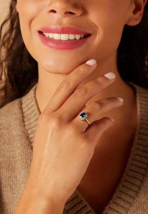 Bague en or avec une pierre précieuse bleue centrale entourée de petites pierres claires. La main du modèle repose sur son visage, mettant en valeur un vernis à ongles clair.