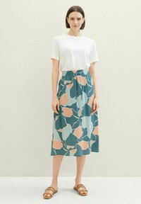 T-shirt blanc léger et jupe midi turquoise avec des motifs floraux abstraits en pêche, bleu et vert. Des sandales décontractées complètent le look.