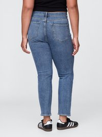Jeans azul de cintura alta com um corte ajustado, apresentando dois bolsos traseiros e uma perna afunilada, combinados com sapatilhas pretas e brancas.