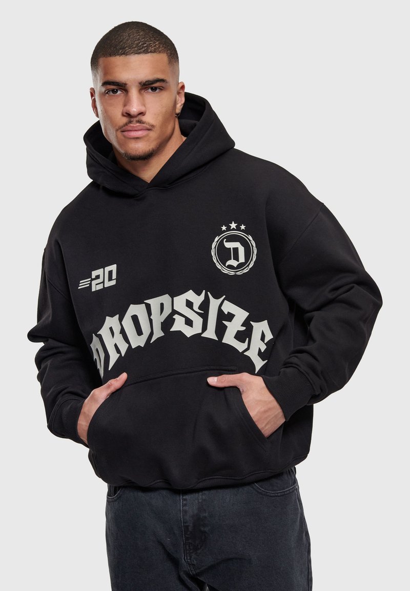 Homme portant un sweat à capuche noir avec le texte « Dropsize », un emblème avec des étoiles, et le détail « 20 », mains dans la poche avant, sur un fond uni.