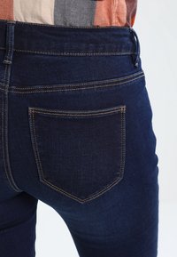Jeans de mezclilla azul oscuro con una silueta ajustada. Características incluyen un solo bolsillo trasero y costuras claras contrastantes. Textura suave.
