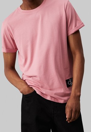 Mand iført en ensfarvet lyserød kortærmet t-shirt med en lille sort logoetiket og sorte bukser, den ene hånd i lommen.