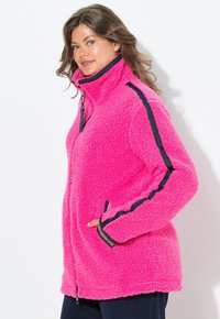 Fleece takki kirkkaan vaaleanpunaisena, jossa on korkea kaulus, vetoketjukiinnitys, sivutaskut ja kontrastiväri mustat raidat hihoissa. Pehmeä tekstuuri.