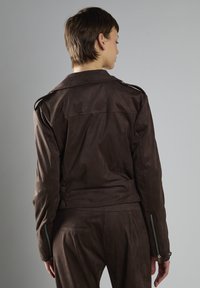 Brun ruskindsjakke med en tætsiddende design, detaljeret syning og lynlåslukninger ved manchetterne. Har skulderepauleter og en glat overflade.