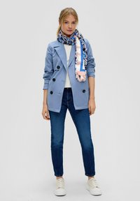 Veste bleu clair à double boutonnage avec des boutons noirs, associée à un haut blanc, un jean bleu foncé et une écharpe à motif bleu et rose. Baskets blanches.