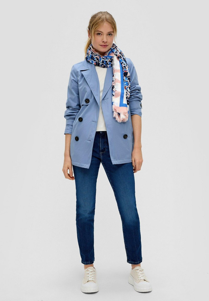 Veste bleu clair à double boutonnage avec des boutons noirs, associée à un haut blanc, un jean bleu foncé et une écharpe à motif bleu et rose. Baskets blanches.