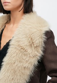 Καφέ σακιέ με ντεμοντέ καφέ σουέντ και φαρδιά, απαλή μπεζ τεχνητή γούνα στον γιακά. Φοράει μαύρο ριμπ τοπ από κάτω και προσφέρει κοντινή ματιά στον λαιμό.