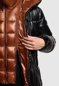 Svart vadderad jacka med glänsande yta, utrustad med en brun quiltad innerfoder och dubbla dragkedjor. En hand syns nära öppningen och visar kontrast i texturen.