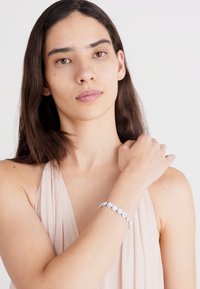 Bracelet en argent avec des accents circulaires et en forme de goutte d'eau. Finition lisse, sertie de pierres précieuses transparentes, porté sur un poignet à la peau claire.