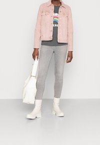 Chaqueta de mezclilla rosa claro sobre una camiseta gris con un gráfico de arcoíris, combinada con jeans skinny grises y botas blancas hasta el tobillo, sosteniendo un bolso blanco.