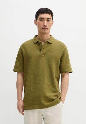 Mężczyzna ubrany w oliwkową, teksturowaną koszulkę polo z krótkim rękawem i jasnobeżowe spodnie, stojący z jedną ręką w kieszeni na jednolitym tle.