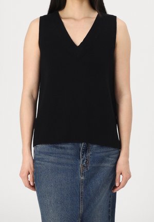Femme portant un haut noir sans manches à col en V associé à une jupe en jean bleue, debout devant un fond clair uni.