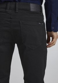 Svarte denimjeans med en glatt tekstur, fem lommer og subtile sømdetaljer. Har et merket svart lærpatch på livet.