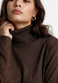 Pull en col roulé marron en maille douce et texturée avec un col côtelé. Le mannequin porte des boucles d'oreilles en or et ses cheveux légèrement froissés sont visibles.
