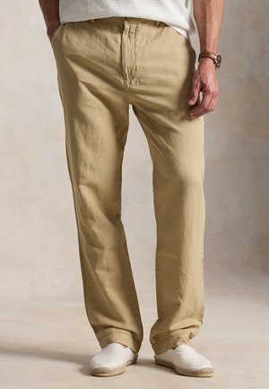 Mężczyzna w luźnych beżowych bawełnianych spodniach, białych butach wsuwanych, białej koszuli i brązowym skórzanym zegarku stoi ze swobodnie opuszczonymi rękami.