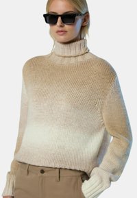 Chunky Strickpullover in beigem Ombre-Muster mit hohem Rollkragen, gerippten Bündchen und weicher Textur. Kombiniert mit schwarzen Sonnenbrillen.