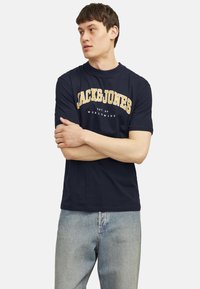 Navy blue cotton t-shirt featuring "JACK&JONES" logo in yellow and white. Short sleeves, crew neck, soft texture. Styled with light blue jeans.