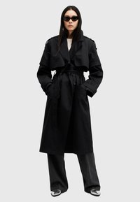 Czarny płaszcz trench z paskiem w talii, szerokimi klapami i kieszeniami po bokach. Model nosi eleganckie czarne okulary przeciwsłoneczne i spodnie w paski.