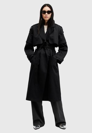 Cappotto nero lungo con vita cintrata, ampie revers e tasche laterali. Il modello indossa occhiali da sole neri eleganti e pantaloni a righe sottili.
