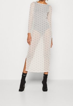 Femme portant une longue robe transparente avec de petits logos Nike partout, des chaussures à talons noires et des chaussettes Nike noires.