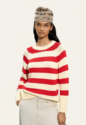 Kobieta ubrana w czerwony i kremowy sweter w paski oraz beżową szydełkowaną czapkę z srebrnymi zdobieniami, stoi z ręką w kieszeni.